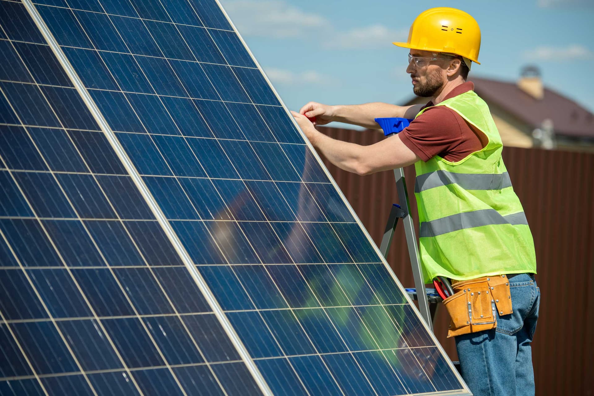 Technician in a hard hat and safety vest fitting a solar panel array from a ladder