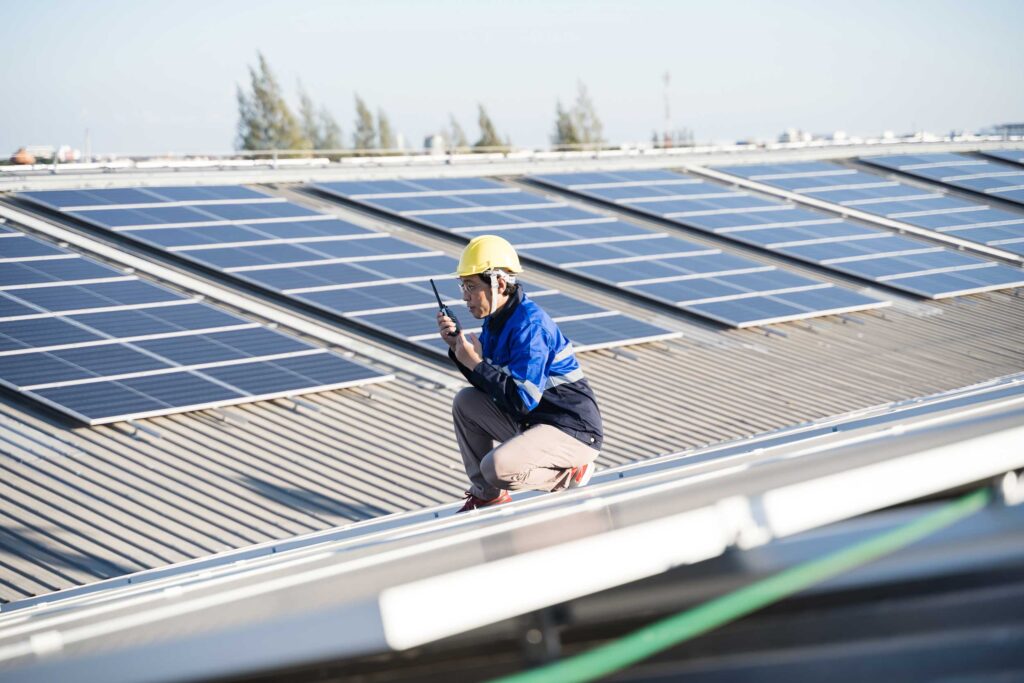A technician in a yellow hard hat and blue jacket is crouched on a large commercial building's roof, inspecting or performing maintenance on an expansive array of solar panels under a clear sky.