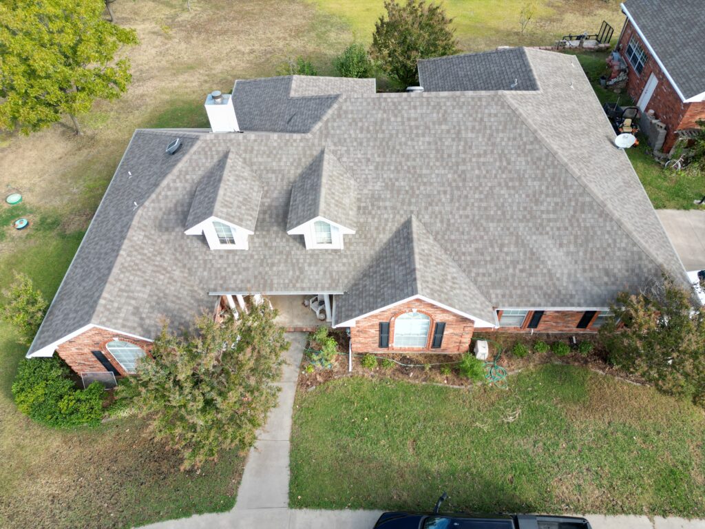 Aerial view of a residential home with a multi-gabled roof, featuring new shingles. The home has a brick facade with white trim, surrounded by a well-kept lawn, walkways, and a driveway with a vehicle parked out front.