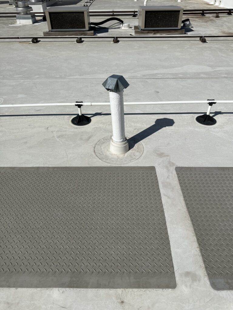 Close-up of a white commercial TPO (thermoplastic polyolefin) roofing system with pitch pan repairs around a vent pipe, and protective walk pads leading to HVAC units in the background, showcasing a neat and professional maintenance setup.