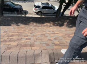 The image shows a view from a rooftop with newly installed brown and tan shingles, with a partial view of a person standing on the roof, and cars parked on the street below, indicating a roof replacement at Hill Street in San Antonio, TX, captured on May 8, 2023.
