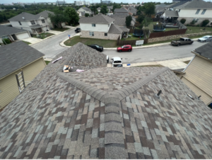 Aerial view of a residential roof replacement in progress at Rhodes Villa, San Antonio TX 78249, showing multicolored shingles and materials laid out, with surrounding homes and vehicles parked along the street.