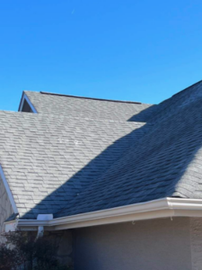 The photo shows a residential home under a clear blue sky with a freshly replaced gray shingle roof, featuring the crisp shadow of an unseen feature casting diagonally across the surface, located in Lantana Ridge, Spring Branch, TX.