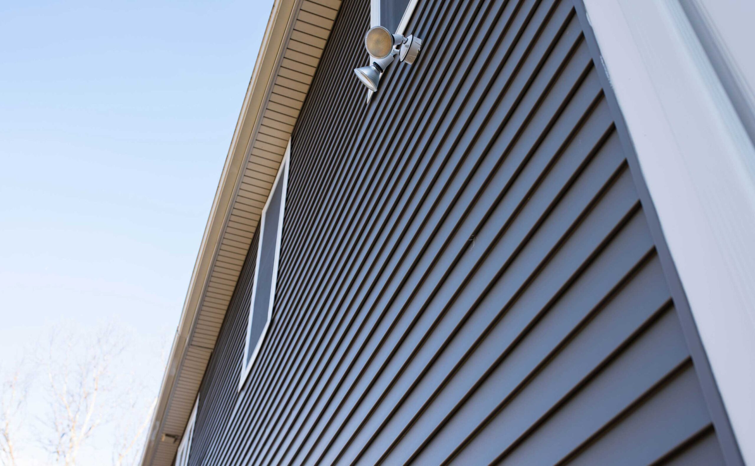 The image captures the architectural details of a house, focusing on the contrasting textures and colors between the beige horizontal siding and the dark vertical siding, accented by a white trim and a security light fixture, under a clear blue sky.