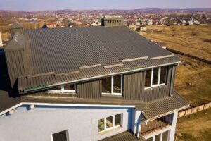 Aerial view of a modern house with a dark metal roof and large windows, located on the outskirts of a suburban area with distant houses and open fields visible in the background.