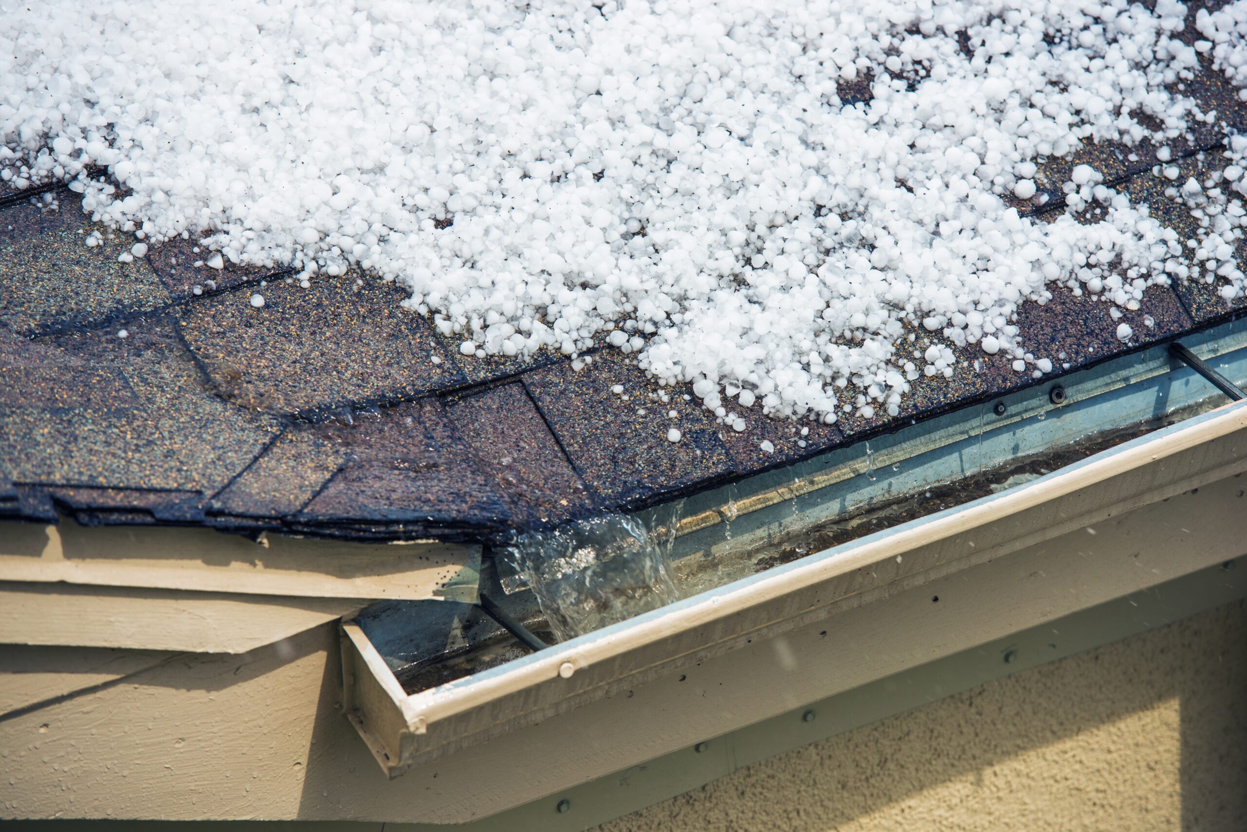 Accumulation of hailstones on a shingle roof, partially filling a gutter, with sunlight casting a glow on the icy white pellets, indicative of a recent hail event.