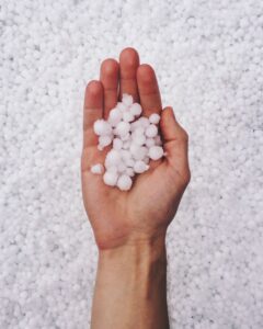 A hand is shown holding a cluster of small, white hailstones against a background completely covered by similar hailstones, showcasing the aftermath of a hailstorm.