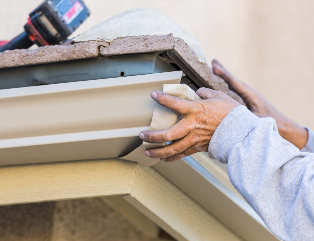 Close-up of a person's hands fitting a beige aluminum rain gutter corner piece onto a house, with a blurred drill in the background on the roof.