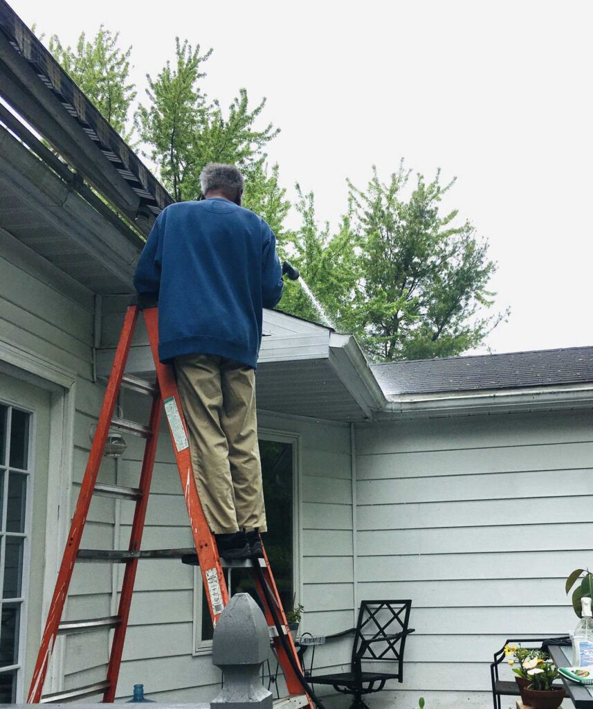 Man on an orange ladder against a house, cleaning the gutter with trees in the background.