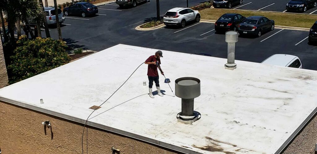 Worker on a flat commercial roof using a pressure washer near a large vent pipe, with a parking lot and cars visible in the background.