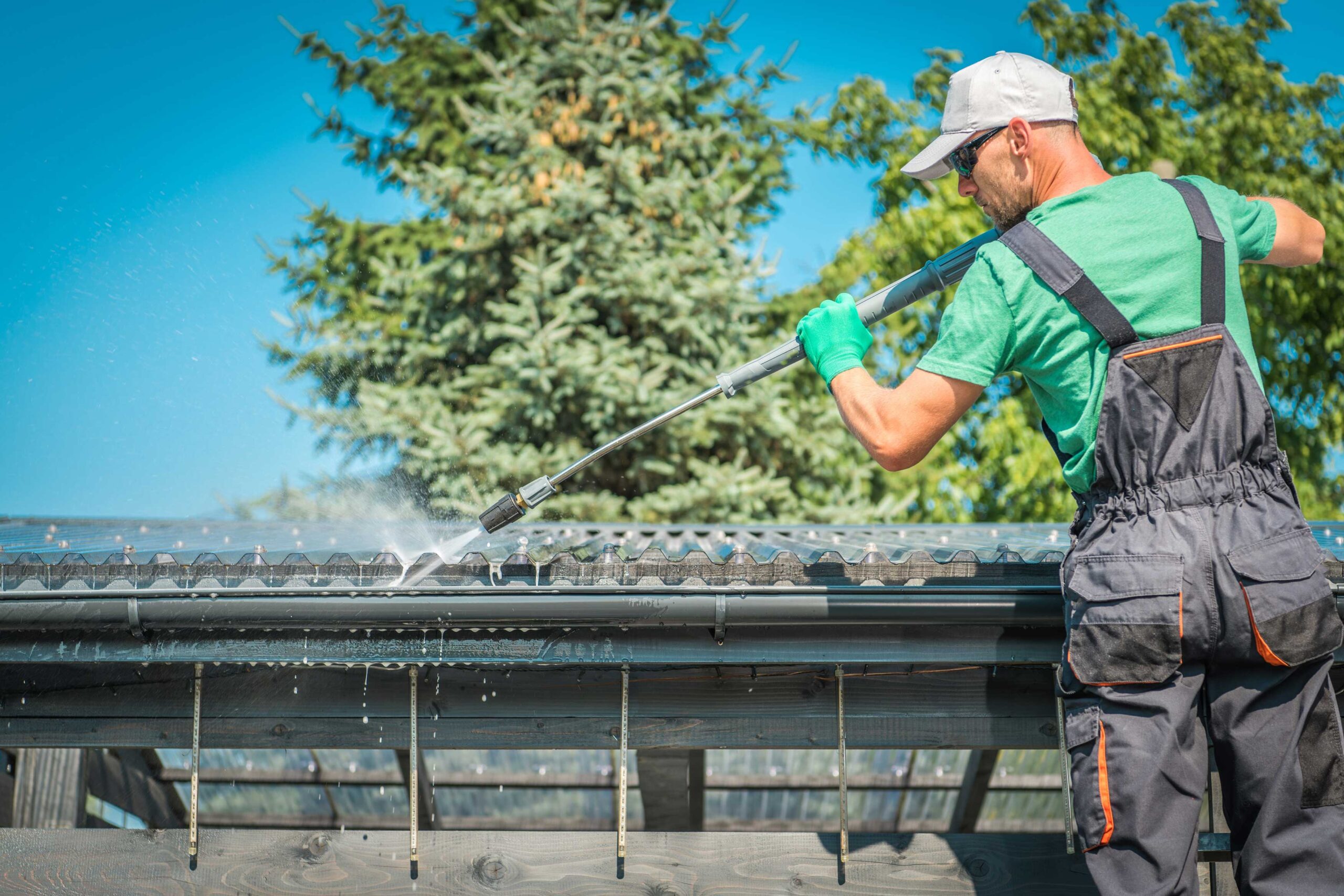 A man in a green t-shirt and dungarees cleaning a roof gutter with a high-pressure water spray, with trees and a clear blue sky in the background.