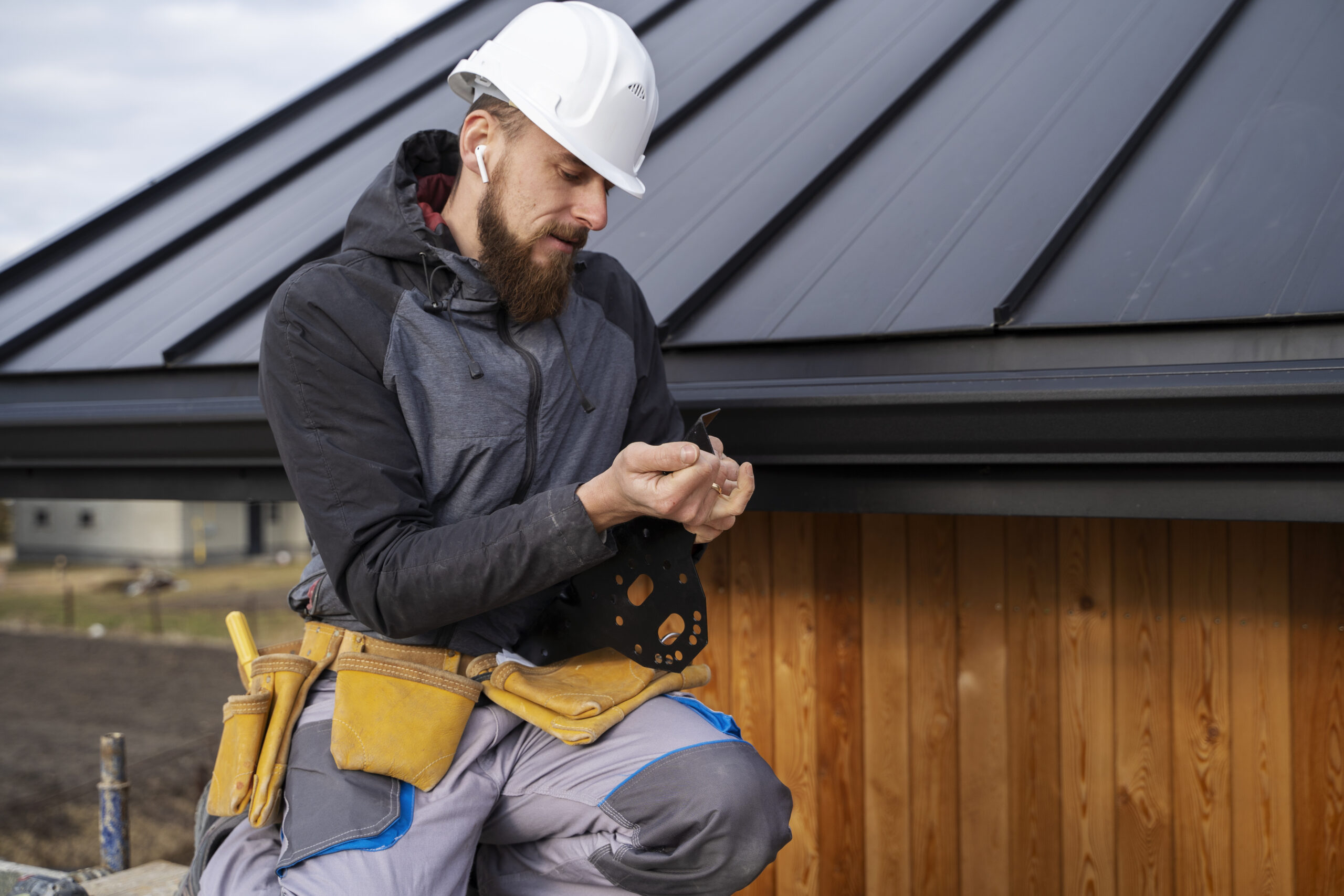 Construction worker with a beard, wearing a white helmet and earbuds, inspecting a small metal object in his hands while sitting on a scaffolding, with a new metal roof and wooden siding in the background.