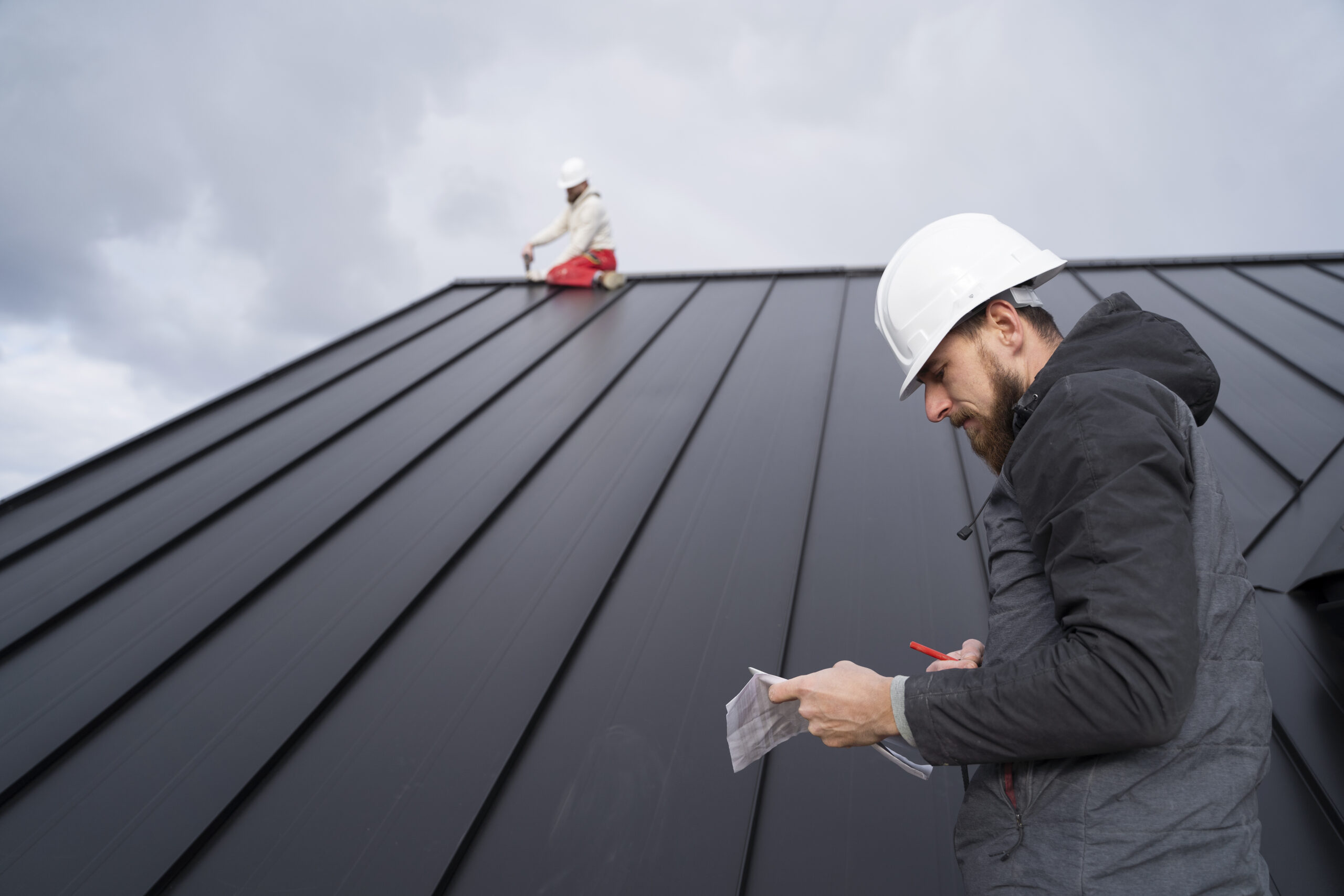 Foreman in a white hard hat and grey jacket writing on a clipboard while conducting a metal roofing inspection, with another inspector in the background examining the roof.