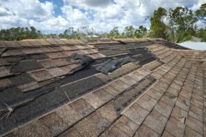 Damaged section of a house roof with missing and torn asphalt shingles, against a backdrop of trees and a clear sky, indicating recent strong weather events or wear.