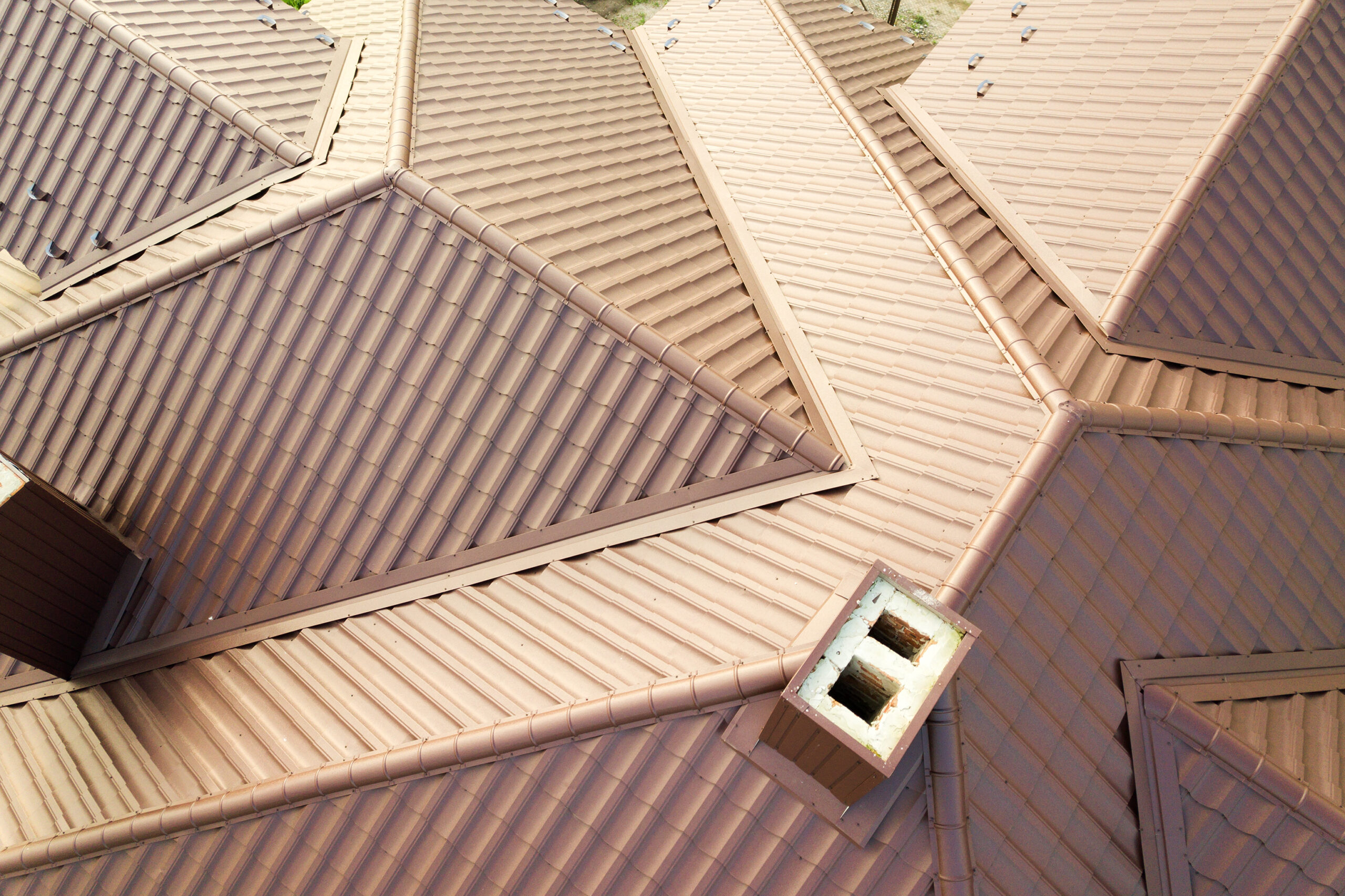 Aerial view of a complex roof structure with multiple ridges and slopes, featuring brown terracotta metal tiles and a single visible chimney.