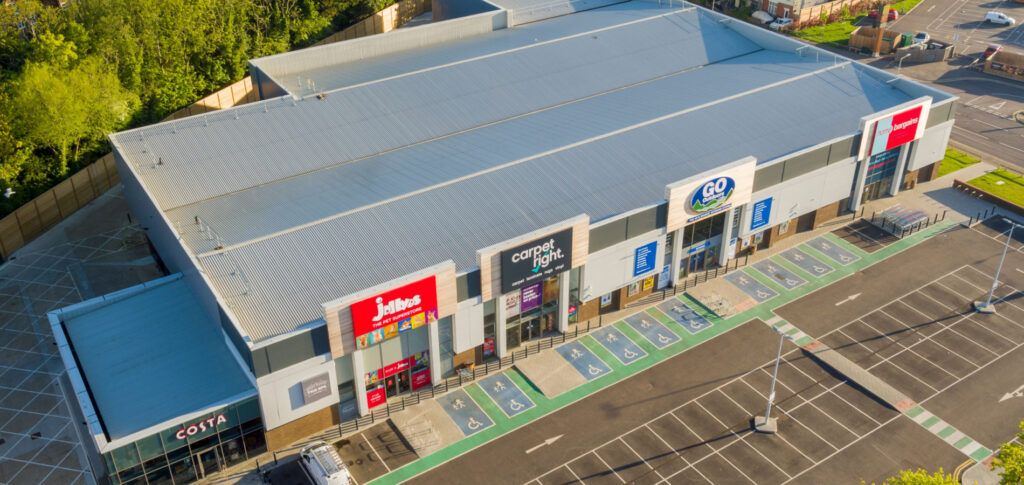 Aerial view of a commercial roofing at a retail park with various storefronts including Costa Coffee, Jollyes, Go Outdoors, and Home Bargains, with a large empty parking lot in the foreground, on a sunny day with clear skies.