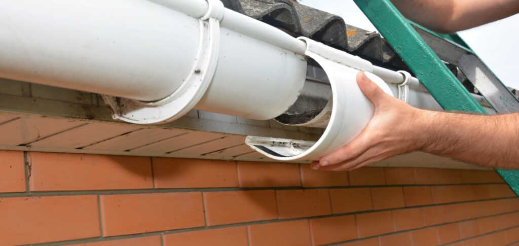 Hands of a person installing a white downspout on a house gutter system, with terracotta brickwork in the background and a tiled roof edge above.
