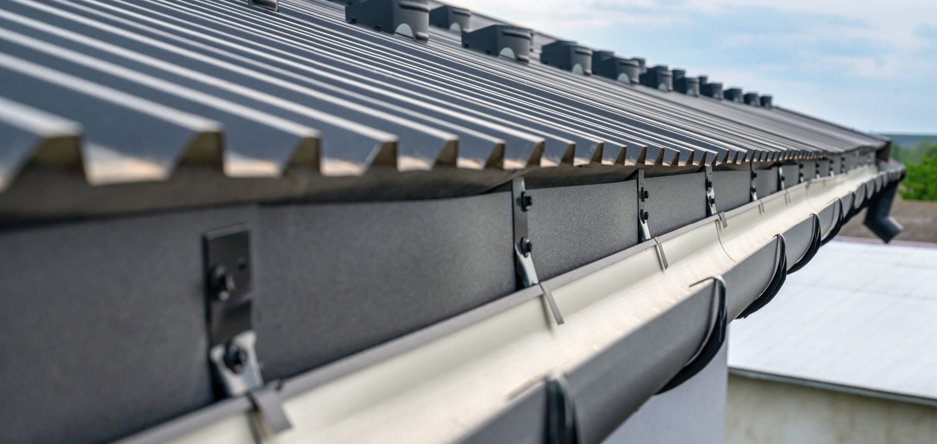 Perspective view of a metal roof with a rain gutter system, showing the texture and pattern of the metal roof panels and the secure mounting of the gutter to the building's fascia.