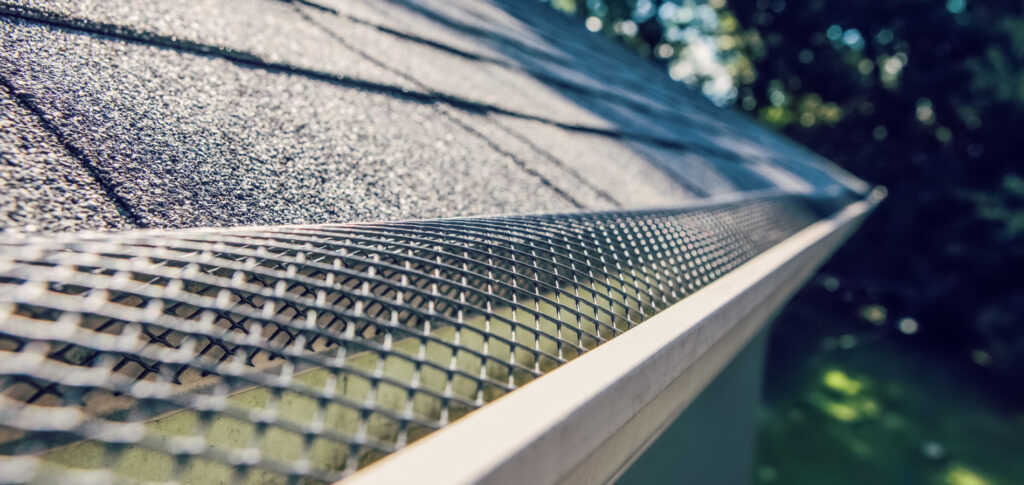 Close-up perspective of a house gutter with a leaf guard mesh installed over it, set against a background of shingle roofing with trees and sky slightly out of focus.