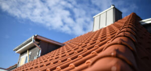 Low angle view of a terracotta tiled roof on a residential home with a chimney and part of the house visible in the background under a blue sky with light clouds.