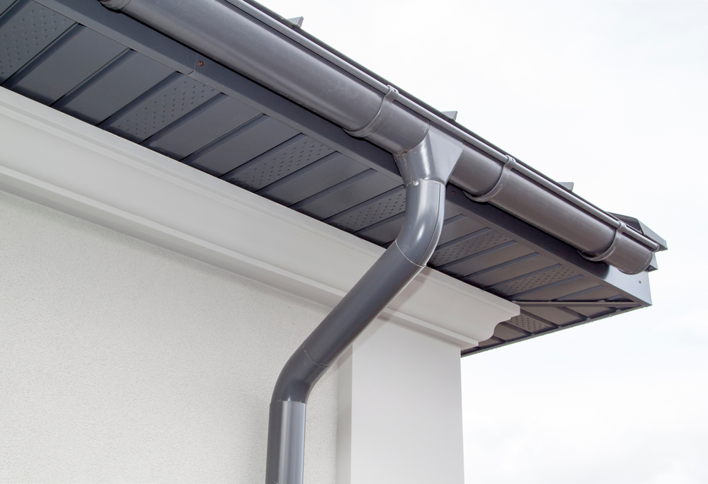 Detail of a house eave with dark grey gutters and downspouts installed against a white soffit, under a roof with grey metal tiles, set against a cloudy sky.
