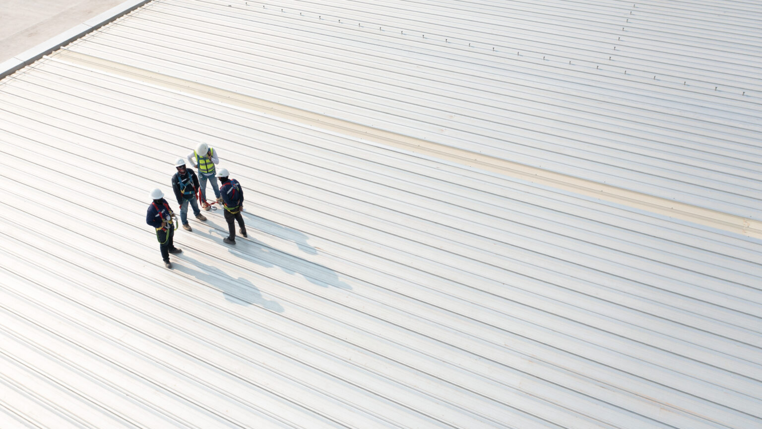 Four construction workers wearing safety harnesses and hard hats on a large white corrugated metal roof, having a discussion.