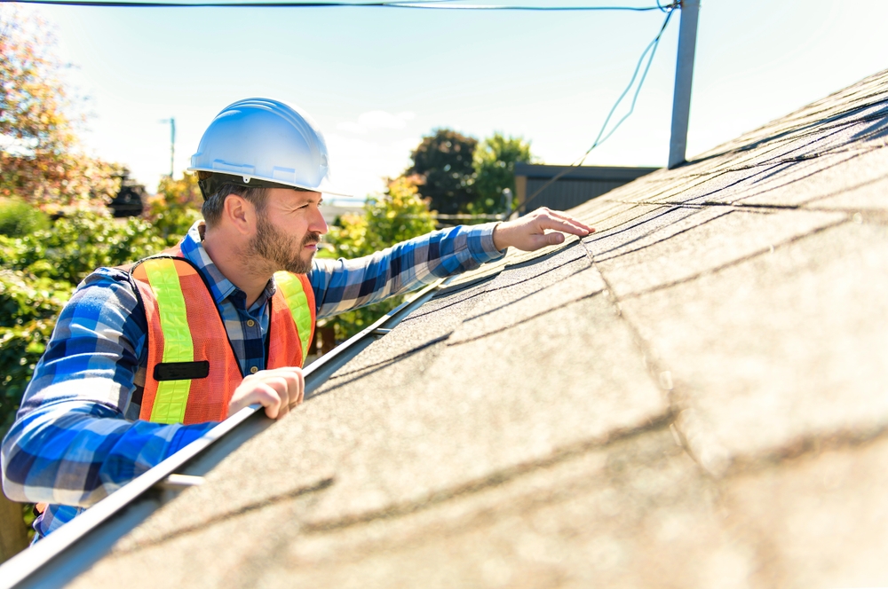 Construction worker in a blue hard hat and high visibility vest inspecting an asphalt shingle roof while holding onto the gutter.