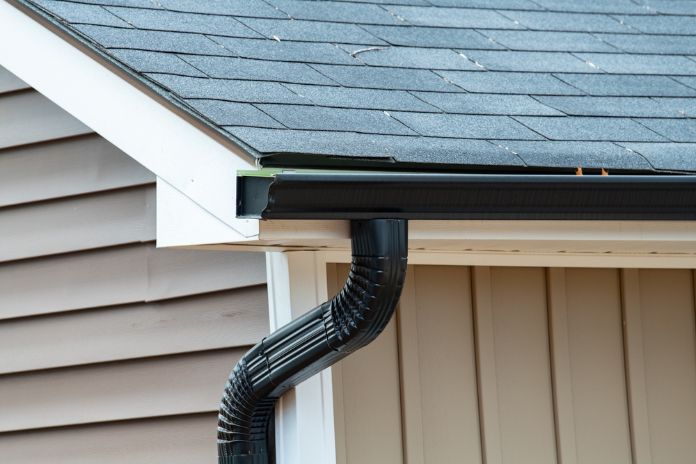 Close-up of a house corner showing asphalt shingles on a sloped roof with a black gutter system and downspout against beige siding.