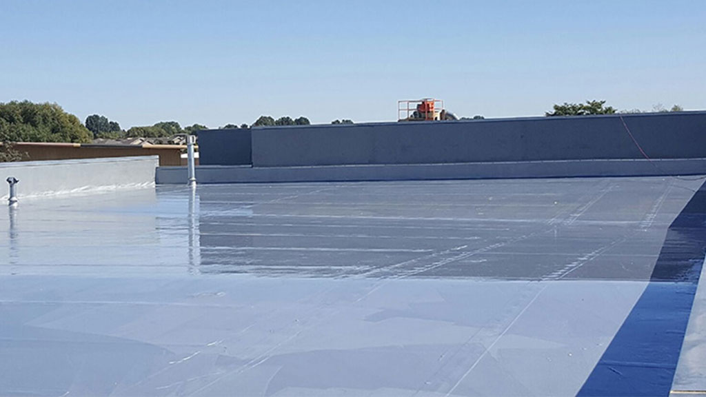 Flat commercial rooftop covered with a grey waterproofing membrane, featuring ventilation pipes and a red safety railing around rooftop equipment, under a clear blue sky.