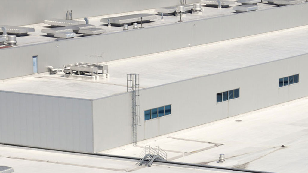 Aerial view of a large industrial building with a flat foam roof, highlighted by air conditioning units, ventilation pipes, and a metal access ladder on the side.