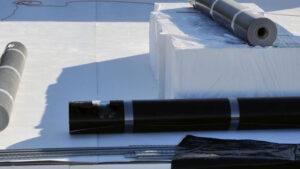 Roofing materials prepared for installation on a flat PVC roof, featuring rolls of black waterproofing membrane, white insulation boards, and additional roofing components.