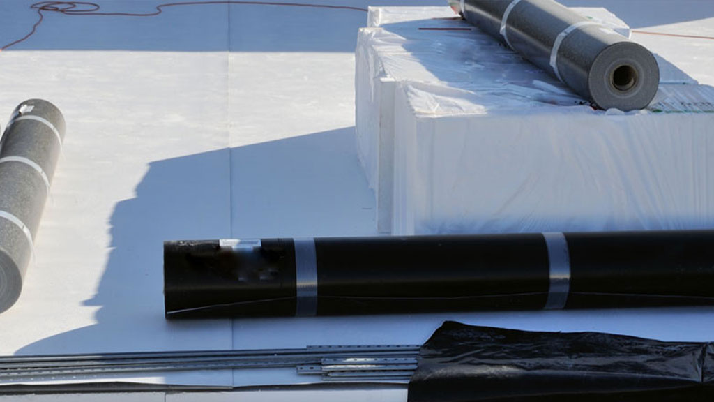 Roofing materials prepared for installation on a flat PVC roof, featuring rolls of black waterproofing membrane, white insulation boards, and additional roofing components.