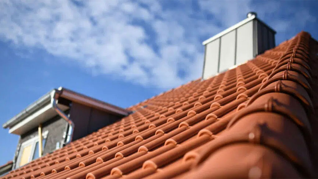 Close-up perspective of a terracotta tiled roof with a blurred background featuring a chimney and the blue sky with scattered clouds.