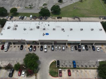 Overhead view of a commercial building with a white flat roof featuring HVAC equipment, next to a parking lot with various vehicles and adjacent green areas.