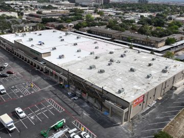 Elevated view of a large commercial building with a flat roof surrounded by a busy parking lot, with an urban landscape in the background.
